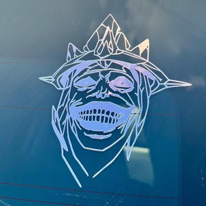 Puede incluir: Una pegatina holográfica que representa una cara estilizada con una estructura similar a una corona. El diseño incluye una boca sonriente y rasgos faciales detallados. La pegatina está diseñada para una ventana de coche o superficie similar.