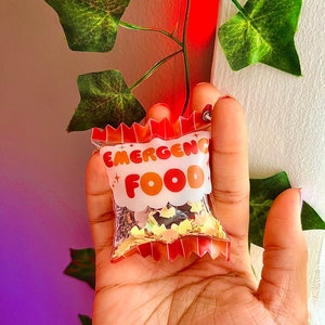 以下が含まれることがあります： 赤い、オレンジ、黄色のデザインで「緊急食」と書かれた透明なビニール袋。金と黒のキラキラが詰まっている。
