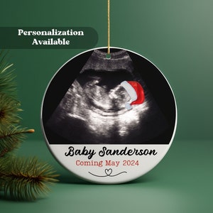 May include: White round Christmas ornament with a black and white ultrasound image of a baby wearing a red Santa hat. The text "Baby Sanderson Coming May 2024" is printed below the image.