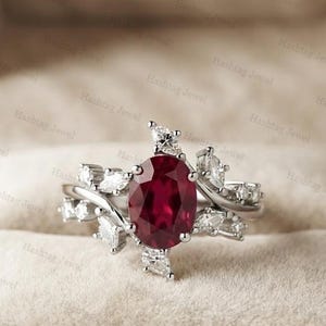 Op de afbeelding: Een zilverkleurige ring met een centrale, ovaal geslepen, dieprode edelsteen. De band is versierd met meerdere kleine, heldere, marquise-geslepen stenen die in een clusterpatroon rondom de middelste steen zijn geplaatst voor een gelaagd effect.
