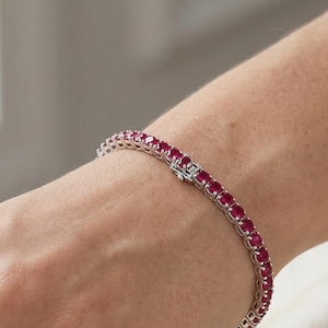 Può includere: Un bracciale tennis caratterizzato da una fila continua di pietre preziose rotonde di un rosso vibrante, incastonate in una fascia di metallo lucido color argento. Il bracciale è dotato di una chiusura a cassetta sicura e flessibile.