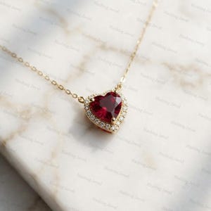 May include: A gold chain necklace featuring a heart-shaped pendant with a deep red gemstone at the center, surrounded by a halo of small white stones. The pendant is attached to a delicate gold link chain that extends upward from the heart.