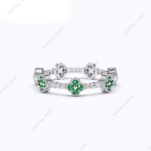 May include: A silver band ring featuring a repeating pattern of four-leaf clover motifs. Each clover is set with four small round green gemstones, and the band is accented with tiny clear white stones between each clover shape.
