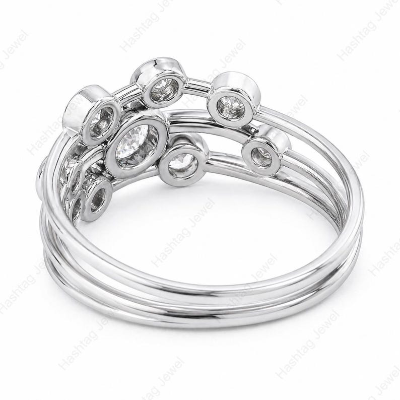 May include: A silver ring with multiple bands and bezel-set diamonds. The ring features a unique design with circular and oval diamond settings, creating a modern and elegant look. The ring is shown against a white background.
