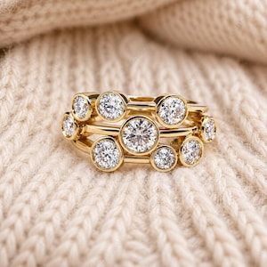 May include: A stack of three gold rings, each adorned with multiple clear, round-cut gemstones. The rings are set against a soft, cream-colored knitted background, highlighting the sparkle of the gems. The rings are a jewelry item.