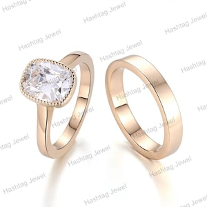 May include: A rose gold ring set consisting of two pieces. The first ring features a large, clear, cushion-cut centre stone with a milgrain-detailed bezel setting. The second ring is a plain, smooth, solid rose gold band with a polished finish.