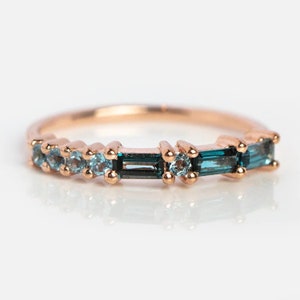 Op de afbeelding: Roségouden ring met afwisselend blauwgroene en lichtblauwe edelstenen. De ring heeft een delicate band en is bezet met een mix van ronde en rechthoekige stenen, wat een uniek en elegant ontwerp creëert.