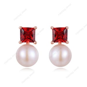 May include: A pair of stud earrings featuring a square, red gemstone atop a white pearl. The earrings have a rose gold setting. The image includes the text "Hashtag Jewel" in the background.