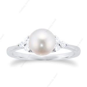 Op de afbeelding: Een zilveren ring met een grote, ronde, witte parel in het midden. Twee groepen kleinere, sprankelende stenen flankeren de parel. De ring heeft een eenvoudig, elegant ontwerp, ideaal voor speciale gelegenheden.