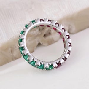 Può includere: Un anello a fascia d'argento con una fila di pietre preziose di colore verde smeraldo e rosso rubino.