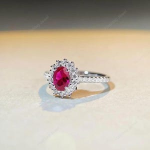 Könnte beinhalten: Ein silberner Ring mit einem ovalen Rubin, umgeben von einem Kranz aus kleinen Diamanten. Das Ringband ist ebenfalls mit Diamanten besetzt, was ein elegantes Design ergibt. Der Ring wird vor einem neutralen Hintergrund präsentiert.