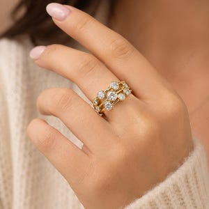 May include: A gold ring with three bands, each adorned with multiple clear, round gemstones. The ring is worn on a hand, showcasing its intricate design and sparkle. The gemstones are set in a way that maximizes their brilliance.