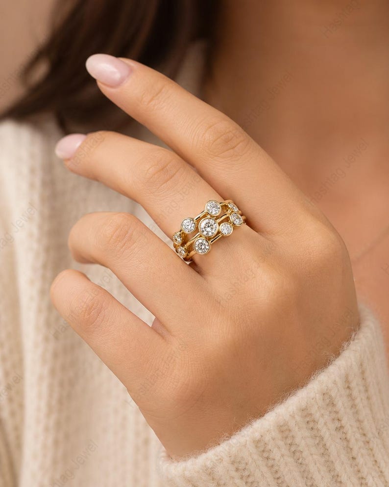 May include: A gold ring with three bands, each adorned with multiple clear, round gemstones. The ring is worn on a hand, showcasing its intricate design and sparkle. The gemstones are set in a way that maximizes their brilliance.