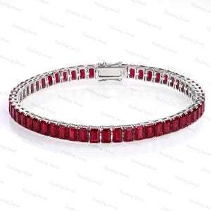May include: A silver-toned tennis bracelet featuring a continuous row of rectangular-cut red gemstones set in a metal frame. The bracelet includes a secure box clasp mechanism with the engraving 14K visible on the metal surface of the closure.