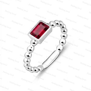 Peut inclure: Une bague argentée ornée d'une pierre précieuse rectangulaire rouge foncé taille émeraude, sertie dans un chaton lisse. L'anneau est composé d'une série de sphères métalliques reliées, se terminant par une section plate et lisse à la base.