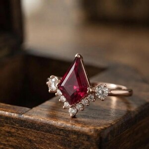 May include: A rose gold ring featuring a central kite-shaped deep red gemstone. The setting is accented with a series of small, round, clear white diamonds arranged in a V-shape along the lower edge of the central stone and on the band sides.