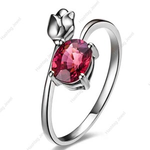 May include: A silver ring with a large, oval-cut, ruby-red gemstone. The ring's design incorporates a stylized flower detail at the top, with the gemstone secured by four prongs. The band curves elegantly, creating a stylish piece of jewellery.