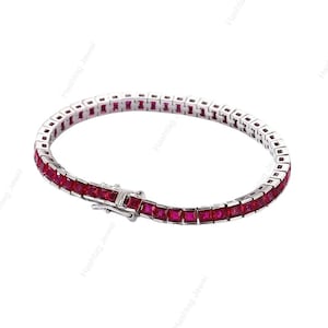 Op de afbeelding: Een zilveren tennisarmband met een rij vierkant geslepen, levendige rode edelstenen. De armband is gesloten en ligt op een witte achtergrond. De merknaam "Hashtag Jewel" is als watermerk in de afbeelding verwerkt.