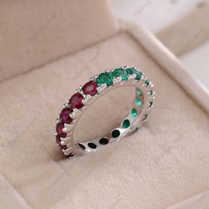 Puede incluir: Un anillo de eternidad plateado con piedras preciosas rojas y verdes alternas. El anillo presenta rubíes y esmeraldas de talla ovalada, creando un contraste vibrante. El anillo se muestra en una caja de joyería de tono neutro.