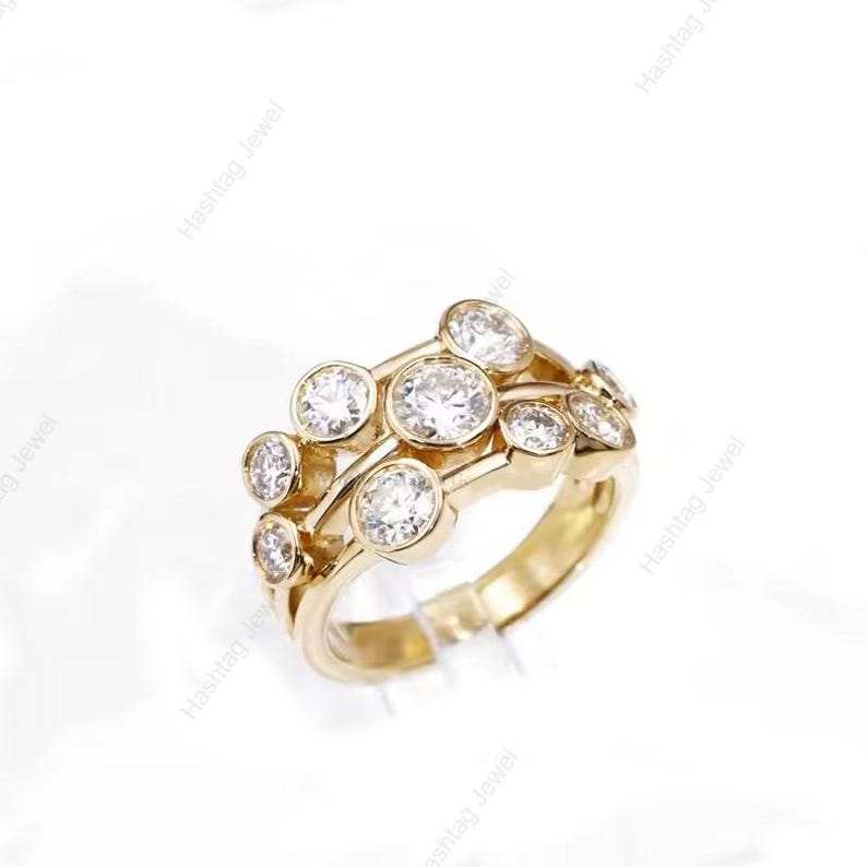 May include: A gold ring featuring multiple clear, round gemstones. The ring has a unique design with the gemstones set in a cluster arrangement. The ring is displayed on a clear stand against a white background, highlighting the jewelry.