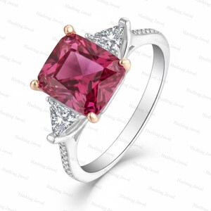 Könnte beinhalten: Ein silberner Ring mit einem großen, kissenförmigen, tiefrosa Edelstein in der Mitte, flankiert von zwei dreieckig geschliffenen weißen Diamanten. Die Ringschiene ist aus poliertem Silber und mit kleinen Pavé-Diamanten besetzt.