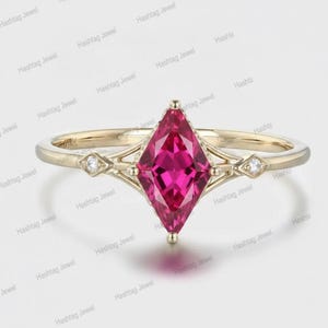 Puede incluir: Un anillo de oro con una piedra preciosa de color rosa vibrante en talla marquesa en el centro. La banda incluye dos pequeñas piedras redondas de acento, una a cada lado del engaste central. El anillo tiene grabado 14K, indicando su pureza.