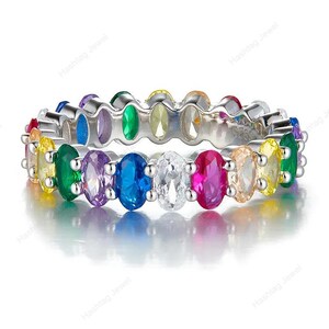 May include: A silver ring featuring a continuous row of oval-cut gemstones in a rainbow of colors. The gemstones include red, orange, yellow, green, blue, purple, and clear stones. The ring is set against a white background.