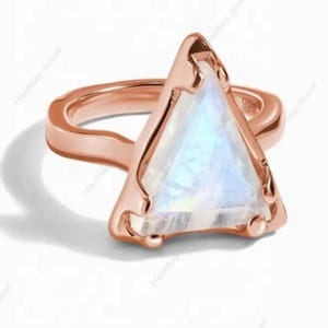 Könnte beinhalten: Roségoldener Ring mit einem dreieckigen, schillernden Mondstein. Der Edelstein hat ein weiches, leuchtendes Aussehen, eingebettet in einen zarten, offenen Rahmen. Der Ring hat ein glattes, poliertes Band und ein modernes, elegantes Design.