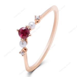 Pode incluir: Um delicado anel de ouro rosa com uma vibrante pedra preciosa vermelha, duas pequenas pérolas e dois minúsculos diamantes. O design do anel é minimalista, com as pedras preciosas agrupadas em um lado da banda.