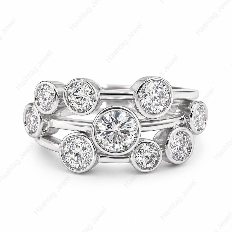 May include: A stack of three silver rings with multiple round, clear gemstones. The rings are set with bezel-set stones of varying sizes, creating a layered effect. The rings are displayed on a white background.