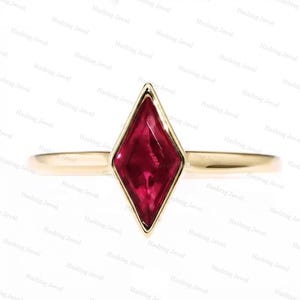 May include: A gold ring featuring a single marquise-cut deep red gemstone set in a vertical orientation. The band is smooth and polished, with a simple metal setting that frames the elongated diamond-shaped stone, creating a clean and focused look.
