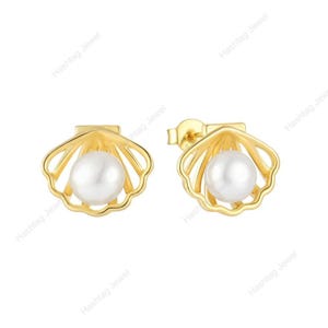 May include: A pair of gold-coloured seashell stud earrings, each featuring a white pearl. The earrings have a detailed, openwork design, with the pearl nestled within the shell's embrace. The earrings are displayed on a white background.