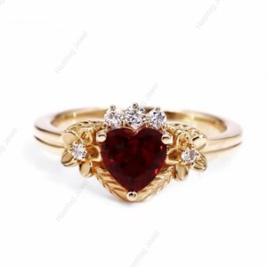 May include: A gold-toned ring featuring a heart-shaped, deep red gemstone at its center. The ring is adorned with small, clear gemstones and floral accents on either side of the heart. The band has a delicate, polished finish.