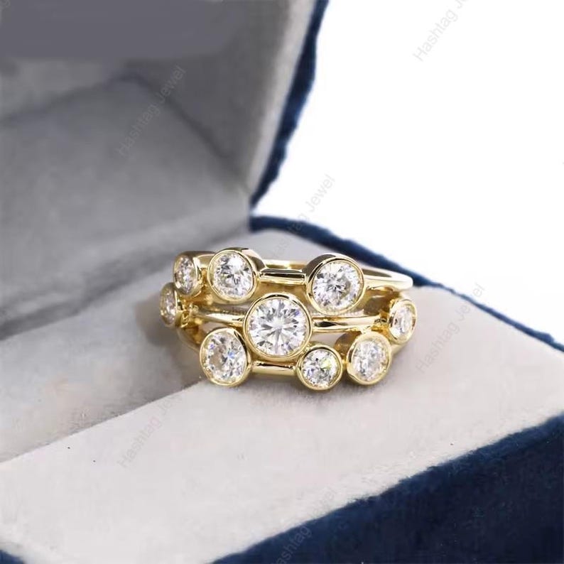 May include: A set of three gold rings with multiple round, clear gemstones. The rings are stacked together, creating a layered effect. The gemstones are set in individual bezels, and the rings are displayed in a blue jewelry box.