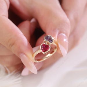May include: A gold ring featuring two heart-shaped gemstones, one ruby red and the other a purplish-blue. The ring is held against a soft, blurred background, highlighting the jewelry's design and color.