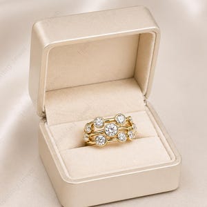 May include: A gold ring with multiple clear, round gemstones is displayed inside a beige jewelry box. The ring features a unique design with the gemstones set in a cluster arrangement. The box is open, revealing the ring against a soft, cream-colored interior.
