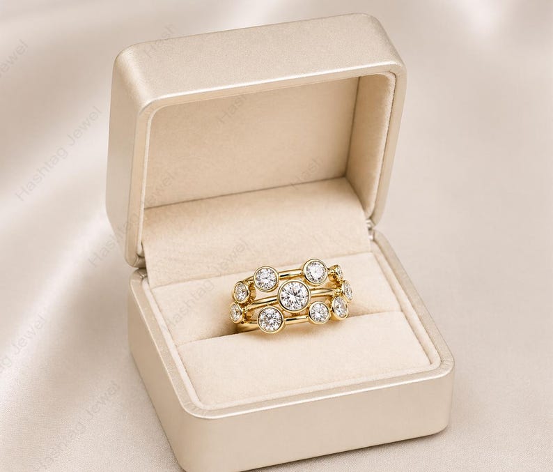 May include: A gold ring with multiple clear, round gemstones is displayed inside a beige jewelry box. The ring features a unique design with the gemstones set in a cluster arrangement. The box is open, revealing the ring against a soft, cream-colored interior.