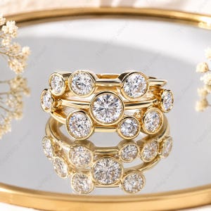May include: A stack of three gold rings with clear, round gemstones. The rings are displayed on a reflective surface, with a gold-colored rim in the background. The gemstones are set in a bezel setting.