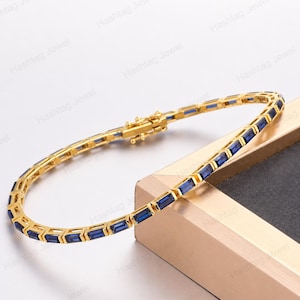 Può includere: Un bracciale dorato con pietre preziose blu rettangolari. Il bracciale ha un design delicato e una chiusura sicura. Le pietre sono uniformemente distanziate. L'immagine è filigranata con "Hashtag Jewel".