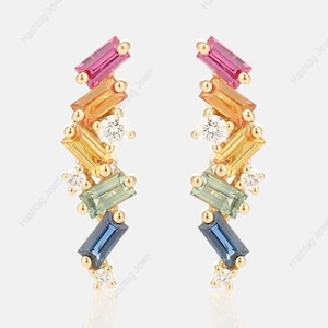 Puede incluir: Un par de pendientes de oro con un diseño colorido. Cada pendiente presenta una serie de piedras preciosas rectangulares en rosa, naranja, amarillo, verde y azul, acentuadas con pequeñas piedras preciosas blancas. Los pendientes están sobre un fondo blanco.