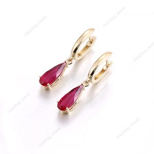 May include: A pair of gold-coloured hoop earrings, each featuring a teardrop-shaped, ruby-red gemstone. The earrings are set against a white backdrop. The text "Hashtag Jewel" is faintly visible in the background.