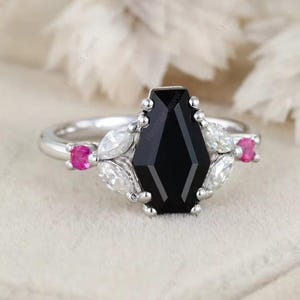 May include: A silver ring featuring a large, black, geometric-cut center stone. The ring is accented with white and pink gemstones, creating a striking contrast. The ring's design is elegant and modern.