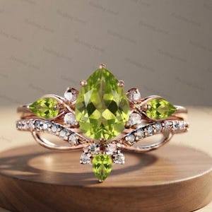 Könnte beinhalten: Ein Ring aus Roségold mit einem großen birnenförmigen Peridot in der Mitte, akzentuiert durch kleinere birnenförmige Peridot-Seitensteine und zahlreiche kleine runde weiße Steine, die entlang der Ringschiene und um den Peridot gefasst sind.