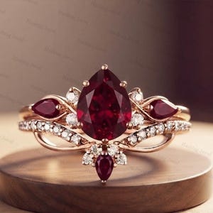 Può includere: Un anello in oro rosa con una grande pietra preziosa rosso scuro a taglio pera come elemento centrale, affiancata da due pietre rosse più piccole a taglio pera. La fascia è decorata con piccole pietre bianche rotonde e altre pietre rosse a pera.