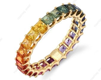 May include: A gold ring featuring a continuous row of square-cut gemstones in a rainbow of colors. The gemstones transition from red to orange, yellow, green, and blue. The ring is set against a white background.