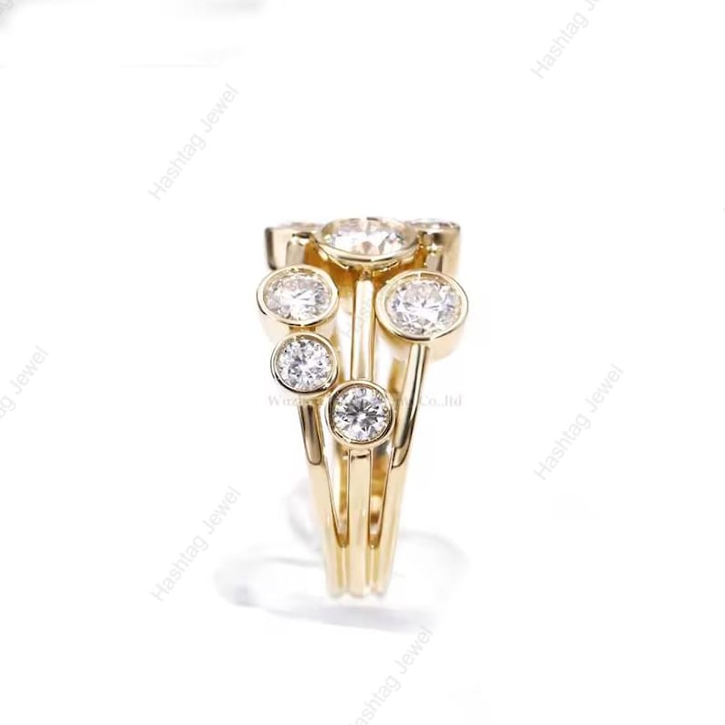 May include: A gold-colored ring with multiple clear, round gemstones. The ring has a unique design with several gemstones set in individual bezels, creating a cluster effect. The ring is set against a white background.