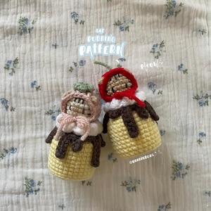 Puede incluir: Dos peluches de gatito pudding de crochet, uno con un sombrero rosa y blanco y el otro con un sombrero rojo y una cereza en la parte superior. Los peluches están sentados sobre un fondo de tela blanca con un patrón floral azul. El texto "Cat Pudding Pattern Meow" está escrito en azul sobre un fondo blanco encima de los peluches.