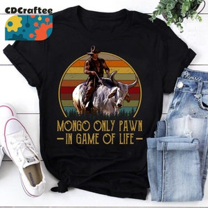 Può includere: Una maglietta nera con una grafica in stile vintage di un cowboy che cavalca un toro bianco. La grafica è su uno sfondo di tramonto con uno schema di colori rosso, arancione e giallo. Il testo "MONGO ONLY PAWN - IN GAME OF LIFE -" è stampato sotto la grafica.