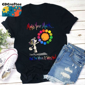 May include: Black t-shirt featuring a colorful graphic of an artist painting a circle of colors. The text "Make Your Mark... And See Where It Takes You!" is printed above and below the graphic. White sneakers and denim shorts are also visible.