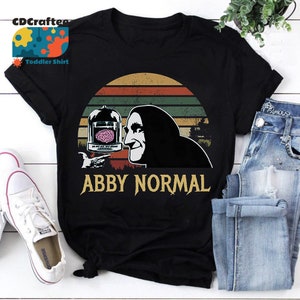 May include: A black t-shirt featuring a vintage-style graphic of a man holding a jar containing a brain. The text "Abby Normal" is printed beneath the graphic.